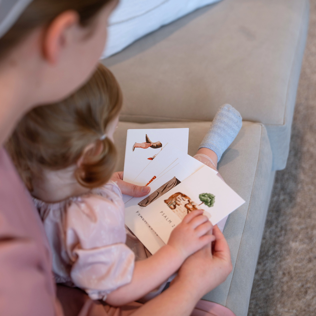 A little Mennonite girl sitting on her mom’s lap on a couch looking at a flashcard with illustrations of Psalm 23